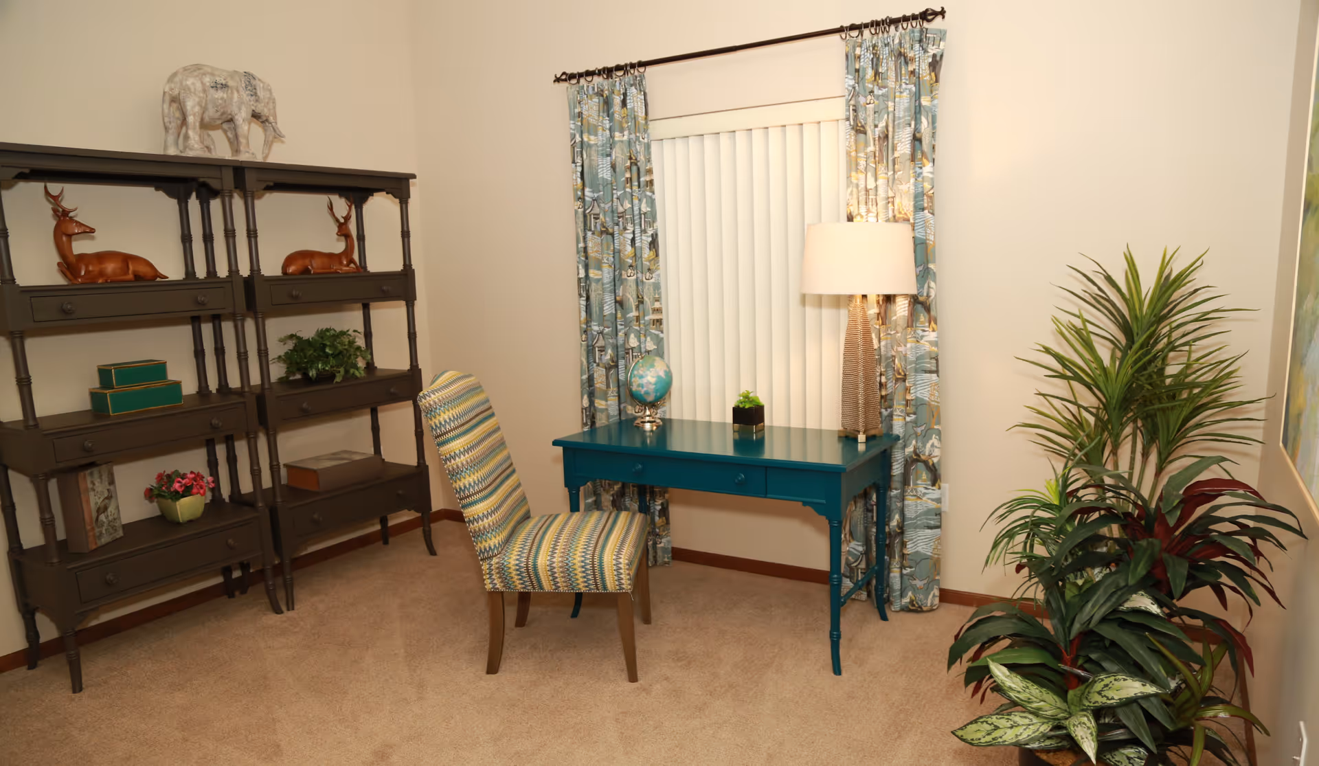 A cozy interior room featuring a teal desk with a globe and a small plant on top, a patterned upholstered chair, a tall lamp, a window with vertical blinds and patterned curtains, a dark wooden shelving unit with decorative items including two deer figurines and a white elephant sculpture, and several green potted plants on the floor.