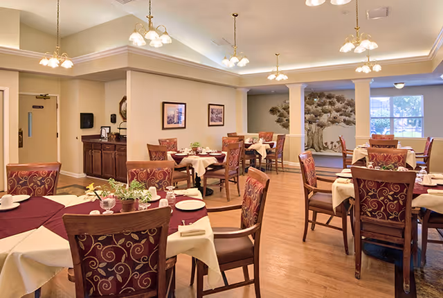 A well-lit dining room with multiple tables covered in white and burgundy tablecloths, each set with plates, cups, and napkins. The chairs have a decorative pattern on the upholstery. The room features wooden flooring, soft overhead lighting, framed pictures on the walls, and a decorative tree sculpture near a window.