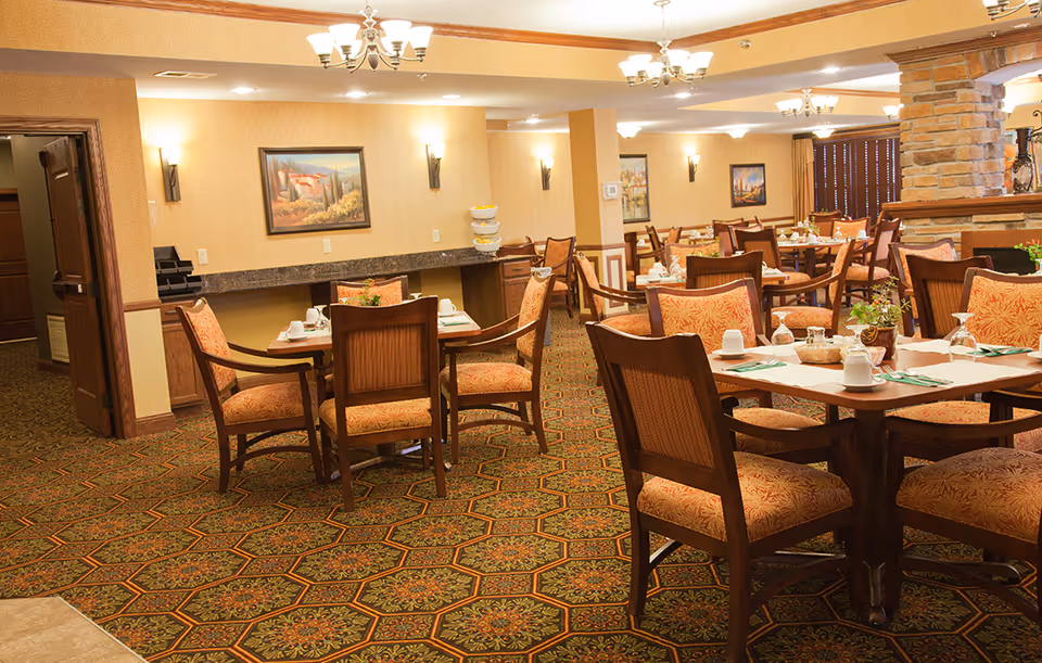Dining area with multiple set tables and upholstered chairs in a senior living facility.