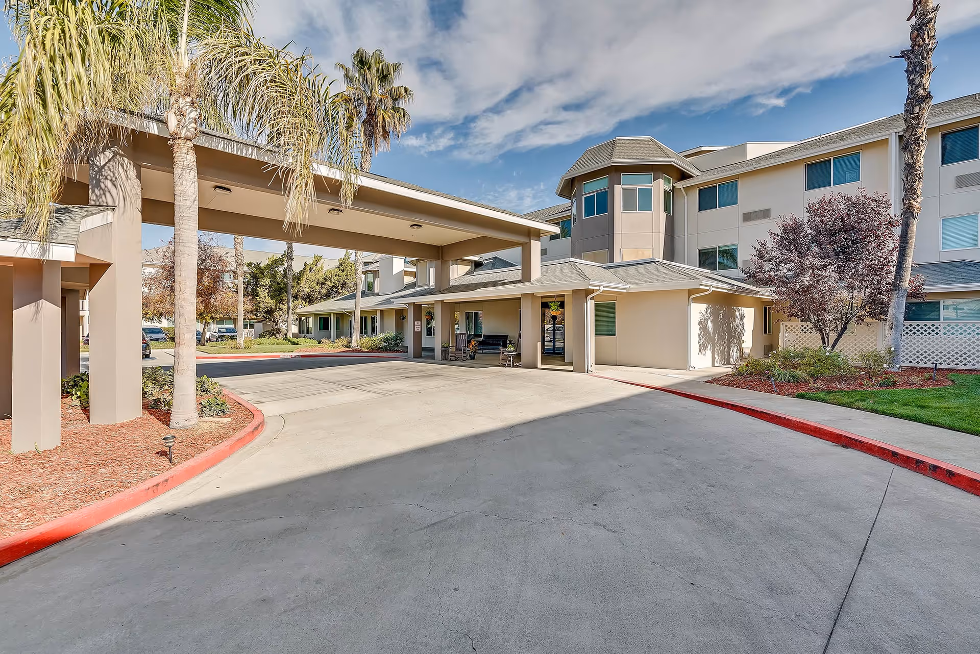 Entrance driveway and covered porte-cochère of a multi-story senior living facility with palm trees and landscaped beds.