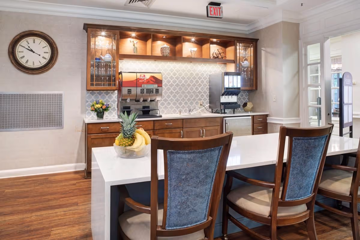 Interior view of a senior living facility's beverage station with wooden cabinets, a coffee dispenser, a soda fountain, a bowl of fruit on a white countertop, and wooden chairs with blue upholstery. A large clock is mounted on the wall, and there is an exit sign above the cabinets.
