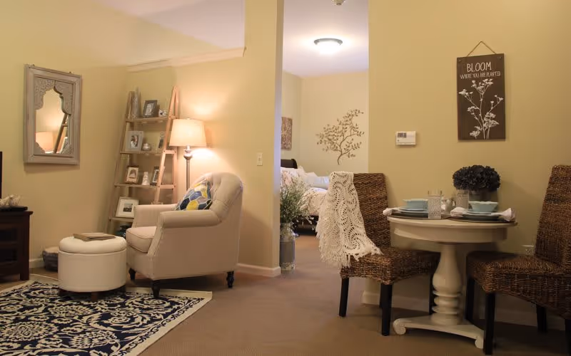 Cozy living area with a beige armchair and ottoman on a patterned rug, a wooden ladder shelf with framed photos and a lamp, a small round dining table set for two with woven chairs, and a view into a bedroom with a bed and wall decor.