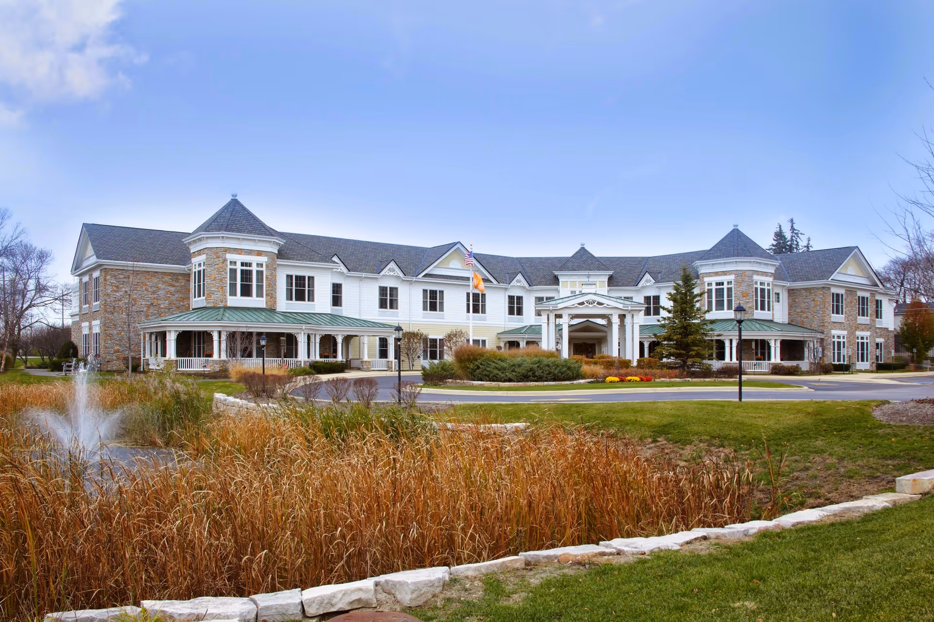 Exterior view of a large two-story senior living facility building with a stone and white siding facade, multiple windows, and a covered entrance. The building is surrounded by landscaped grounds including a pond with a fountain, tall grasses, and a green lawn under a clear blue sky.