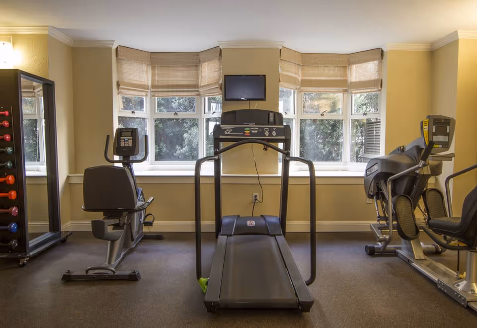 A small fitness room with exercise equipment including a treadmill in the center, a recumbent bike on the left, and an elliptical machine on the right. There is a rack of colorful dumbbells and a full-length mirror on the far left. The room has beige walls, carpeted floor, and a set of windows with bamboo blinds letting in natural light. A small TV is mounted on the wall above the treadmill.