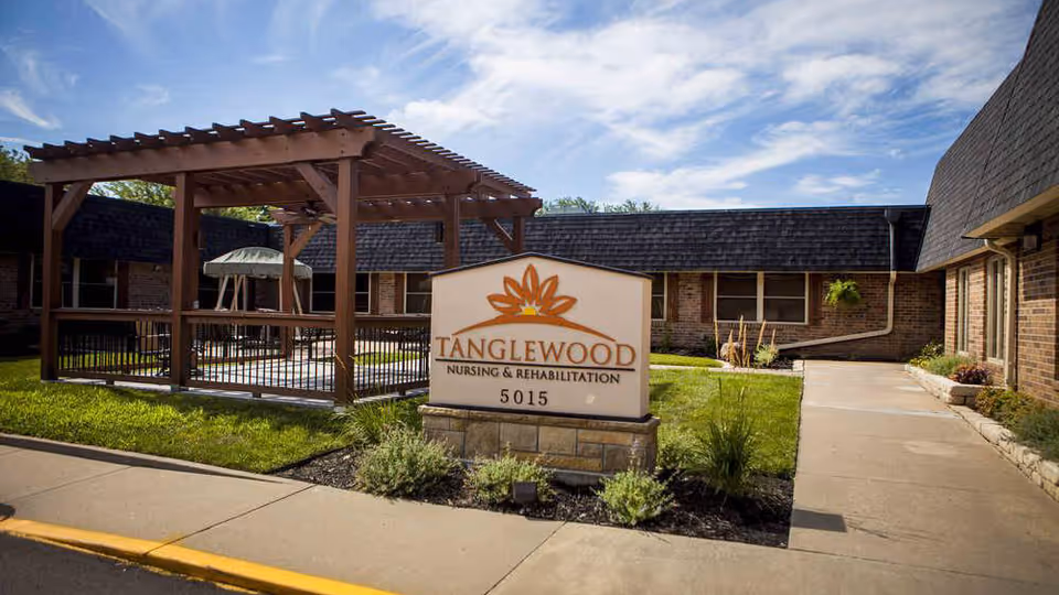 Courtyard with a pergola and a sign reading 'Tanglewood Nursing & Rehabilitation' in front of a single-story brick nursing facility.
