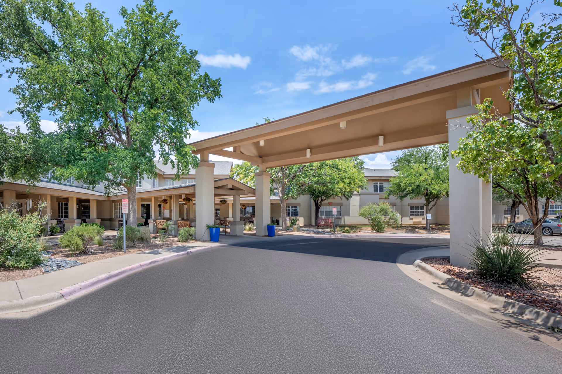 Entrance area of a senior living facility with a covered drop-off zone supported by large columns, surrounded by trees and landscaping under a blue sky with some clouds.