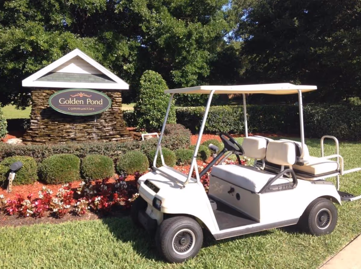 A white golf cart parked on a grassy area next to a landscaped garden with bushes and flowers. Behind the garden is a stone sign with a small roof that reads 'Golden Pond communities'. Trees and greenery are visible in the background.