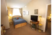 A cozy bedroom with a wooden bed frame and blue bedding, a bedside table with a lamp, a flat-screen TV on a wooden stand, a floor lamp, framed pictures on the beige walls, and a doorway leading to another room.