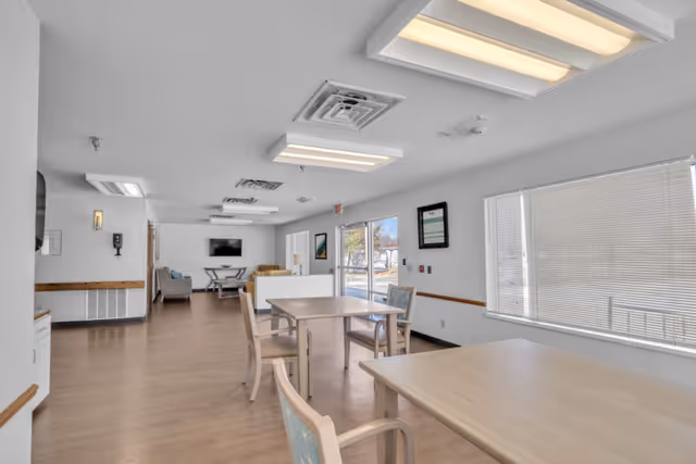 A bright and spacious common area in a nursing retirement home featuring light wood tables and chairs arranged for dining or socializing. The room has large windows with blinds, white walls, and ceiling lights. In the background, there is a seating area with couches and a wall-mounted television.