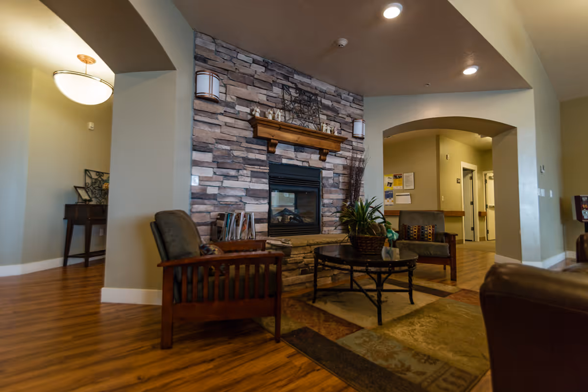 Cozy assisted living common area with a stone fireplace, armchairs, and a round coffee table beneath arched ceilings.