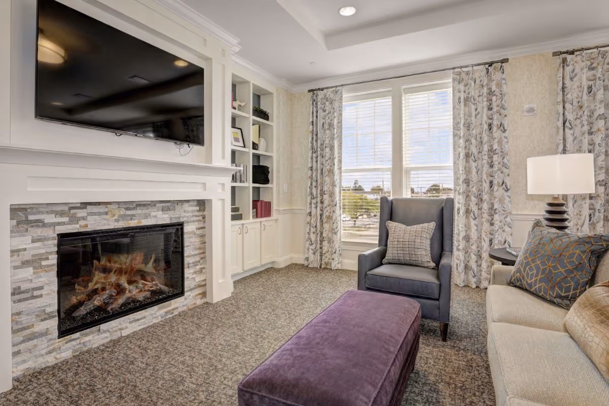 A cozy living room with a modern fireplace beneath a mounted flat-screen TV. The room features a blue armchair with a patterned pillow, a beige sofa with decorative cushions, a purple ottoman, and a side table with a lamp. Large windows with floral curtains allow natural light to fill the space.