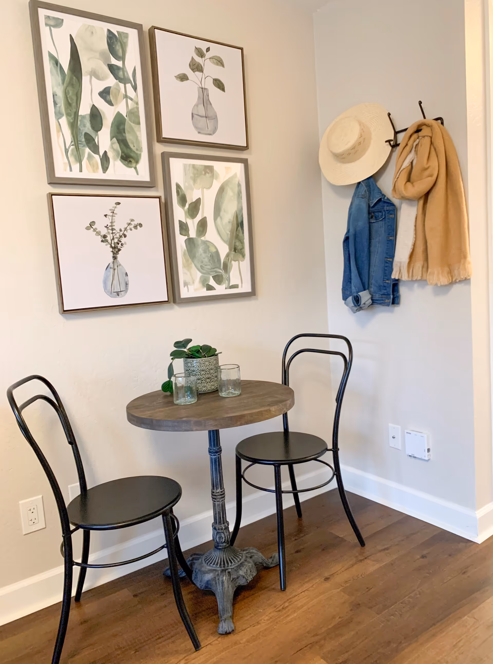 A small round wooden table with a decorative plant and two glasses on top, accompanied by two black metal chairs. On the wall above the table are four framed botanical prints. To the right, a wall-mounted coat rack holds a straw hat, a denim jacket, and a tan scarf. The floor is wooden, and the walls are light-colored.