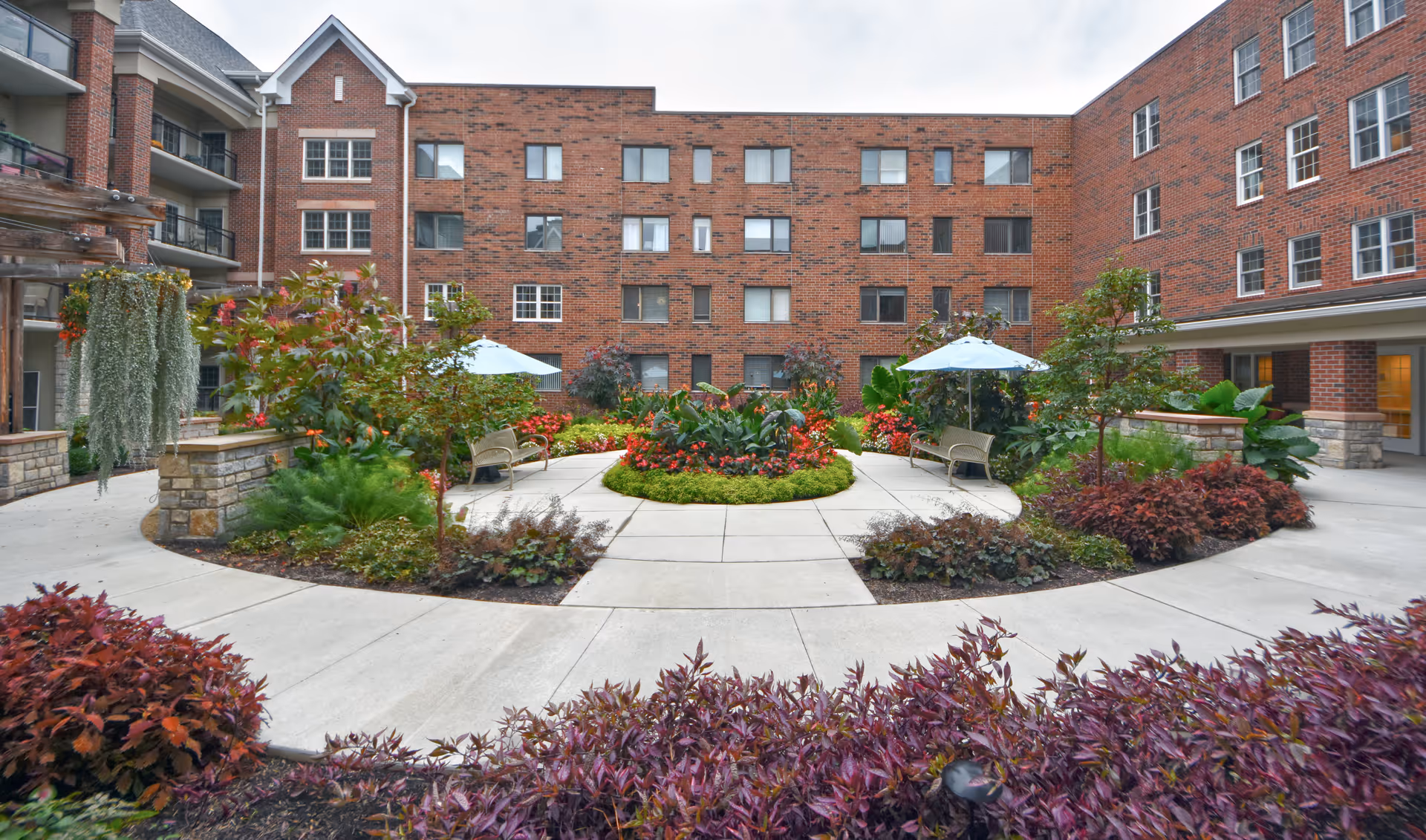 Outdoor courtyard area of a senior living facility with a circular concrete walkway surrounding a landscaped garden bed filled with various plants and flowers. There are benches with umbrellas for shade and a multi-story brick building in the background.