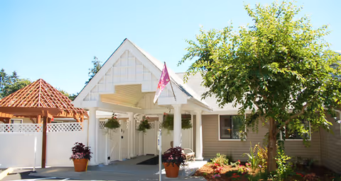Exterior view of a senior living facility entrance with a covered porch supported by white columns, a flag hanging from the roof, potted plants, a wooden gazebo structure, and landscaping with trees and shrubs under a clear blue sky.