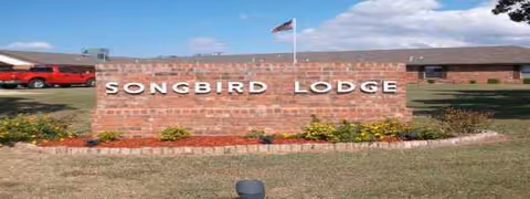Brick sign reading Songbird Lodge with lawn, flowers, flag, and blue sky.