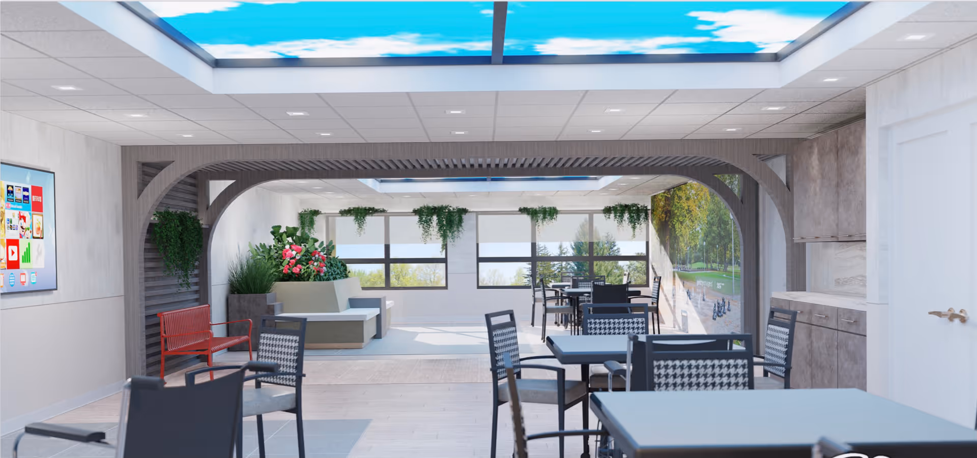 A bright and modern common area with multiple tables and chairs arranged for seating. The ceiling features a large light panel displaying a blue sky with clouds. There are plants and greenery hanging and placed around the room, with large windows letting in natural light. A red bench and a wall-mounted screen are visible on the left side, and a kitchenette area with cabinets and a countertop is on the right.