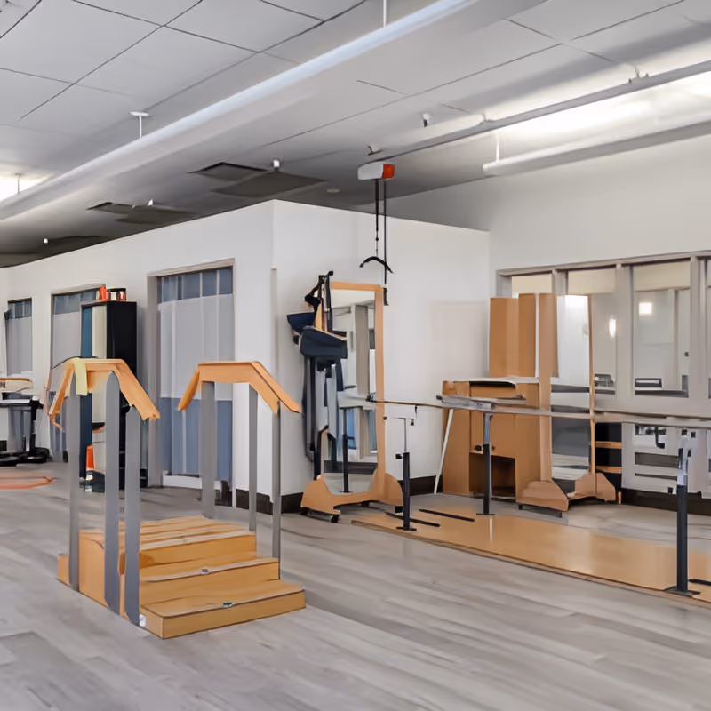 A rehabilitation or physical therapy room with wooden stairs and handrails for walking exercises, a large mirror on the wall, and parallel bars for balance training. The room has light-colored flooring and white walls with some windows and a ceiling with exposed pipes.