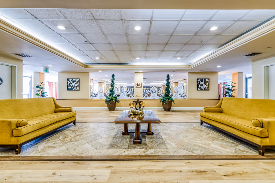 A spacious and well-lit common area with two mustard yellow sofas facing each other across a wooden coffee table. The floor has a combination of wood and tile, and there are decorative plants and framed artwork on the walls. The ceiling has recessed lighting and a drop ceiling design.