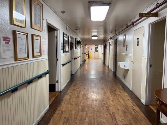 A long, well-lit hallway in a senior living facility with wooden flooring and handrails on both sides. Several doors line the corridor, with framed certificates and pictures hanging on the walls. A small wall-mounted sink is visible on the right side, and a sign indicating an office is also present. The hallway extends into the distance with a caution wet floor sign visible at the far end.