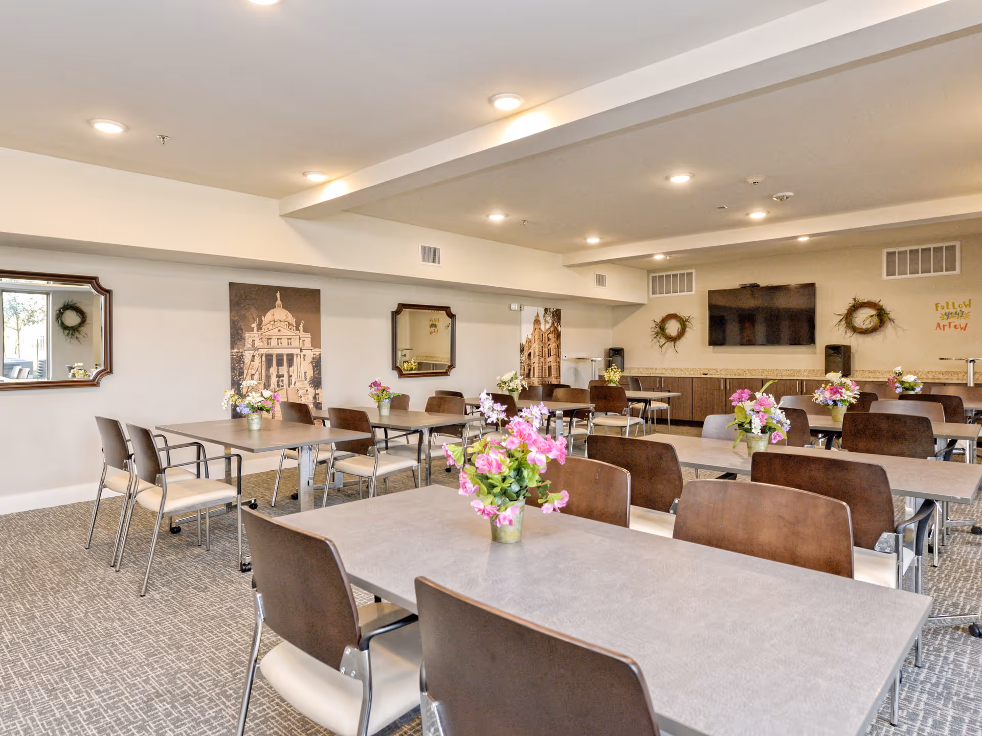 Bright communal dining/activity room with multiple tables and chairs, floral centerpieces, a wall-mounted TV and decorative mirrors.