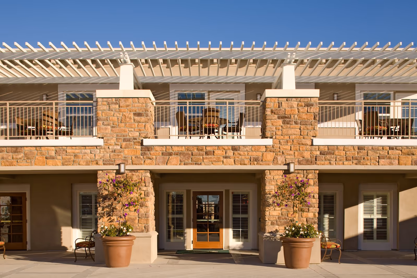 Exterior view of a senior living facility building with stone pillars and beige walls. The upper level features a balcony with a pergola roof and outdoor seating. Large potted plants with flowers are placed on the ground level near the entrance doors.