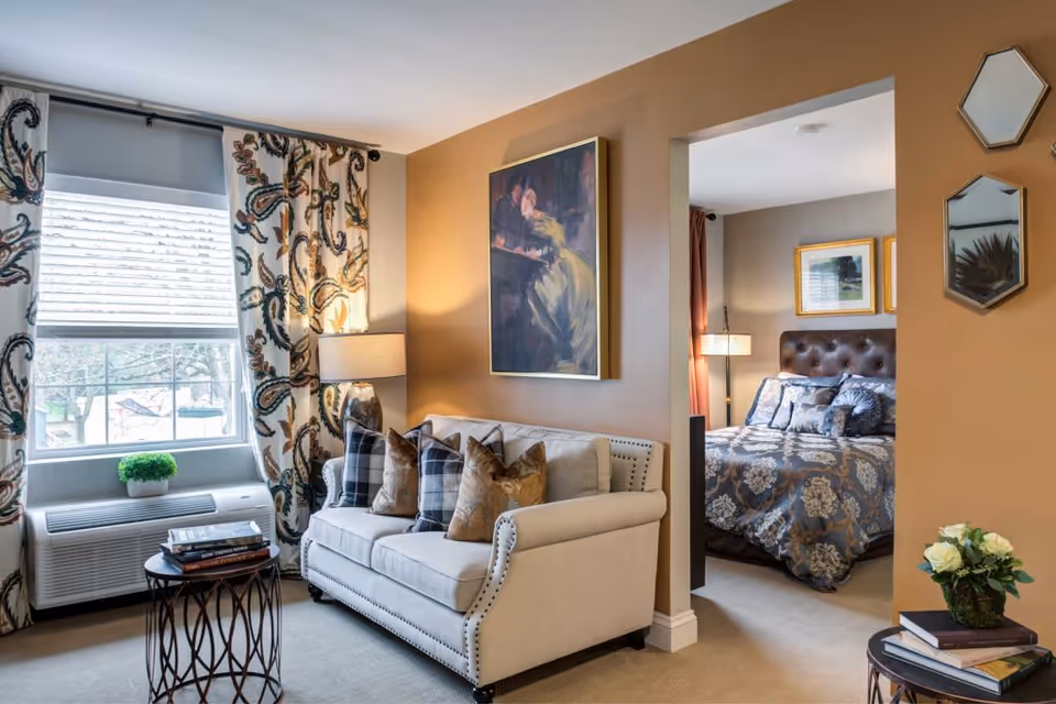 Well-decorated living room with a loveseat, patterned curtains, side tables and a view into an adjacent bedroom.