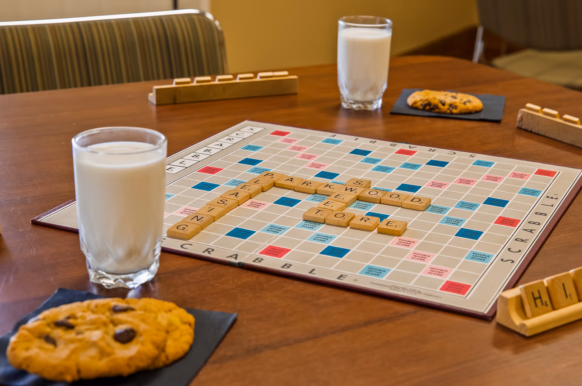 A Scrabble board on a wooden table with the words 'PARKWOOD', 'LIVING', 'EASTING', and 'STORE' formed with letter tiles. There are two glasses of milk and two chocolate chip cookies on black napkins on the table. Scrabble tile racks with letters are also visible around the board.