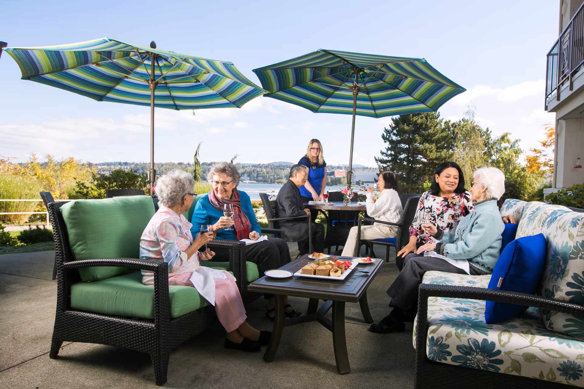 A group of elderly people and a caregiver enjoying a sunny day on an outdoor patio with green and blue striped umbrellas. They are sitting on cushioned wicker furniture, chatting and drinking beverages, with a table in the center holding plates of snacks. Trees and a body of water are visible in the background.