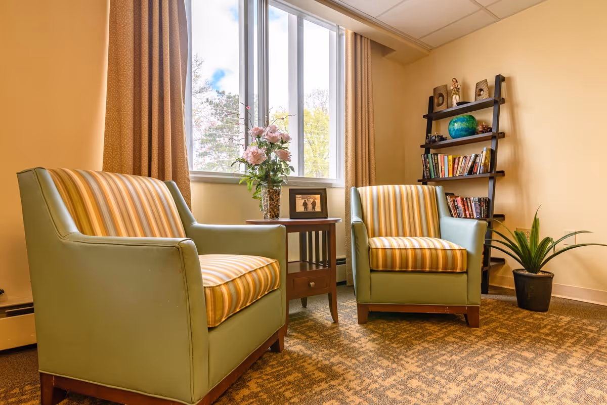 A cozy sitting area with two green armchairs featuring striped cushions, a small wooden side table with a vase of pink flowers and a framed photo, a window with beige curtains letting in natural light, a black ladder-style bookshelf with books and decorative items, and a potted plant on the floor.