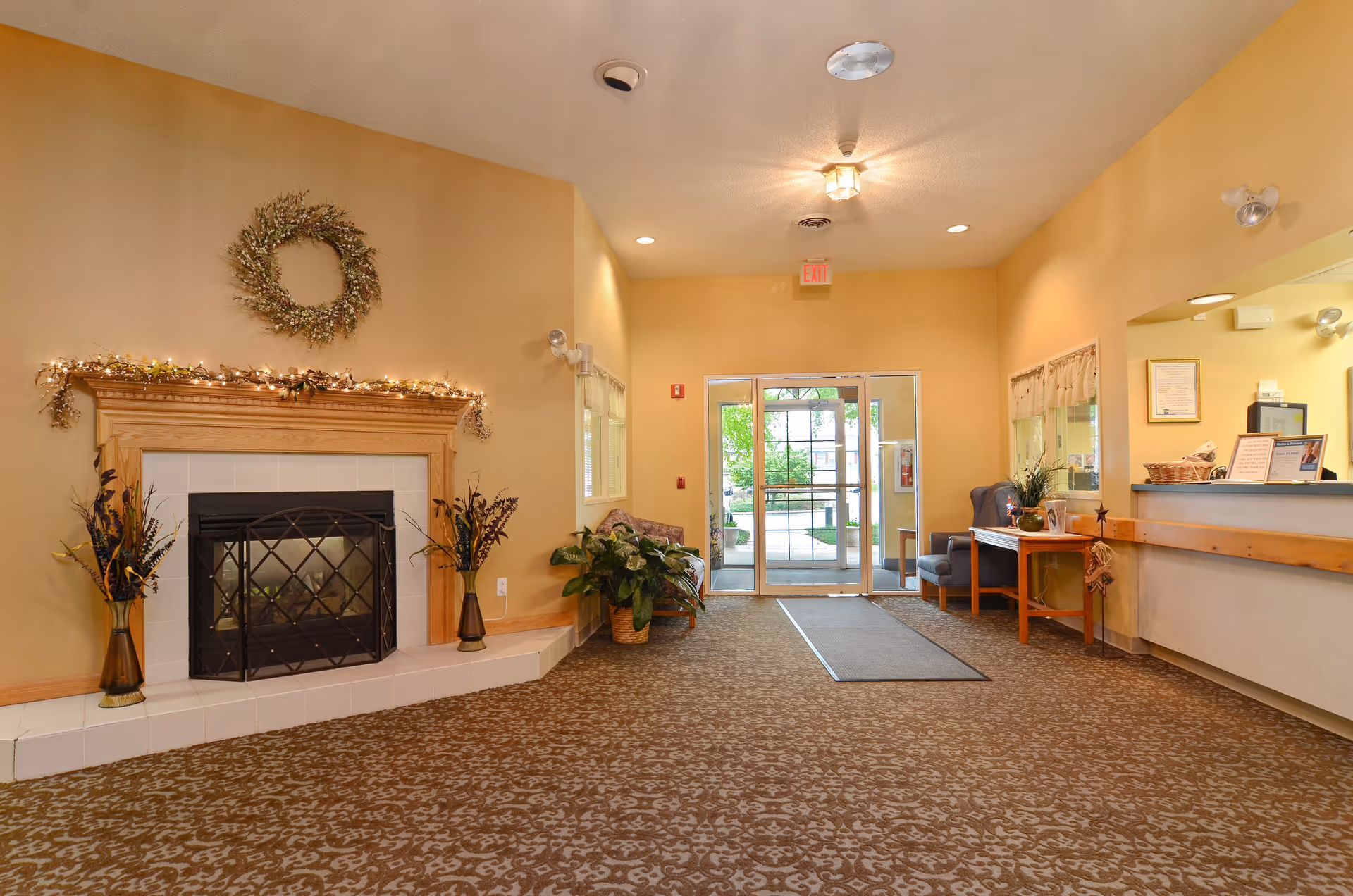 Interior view of a senior living facility lobby with a fireplace decorated with a wreath and string lights, two vases with plants on either side of the fireplace, a patterned carpet, a seating area with chairs and a table, and a reception desk on the right. Glass double doors lead outside, allowing natural light to enter.