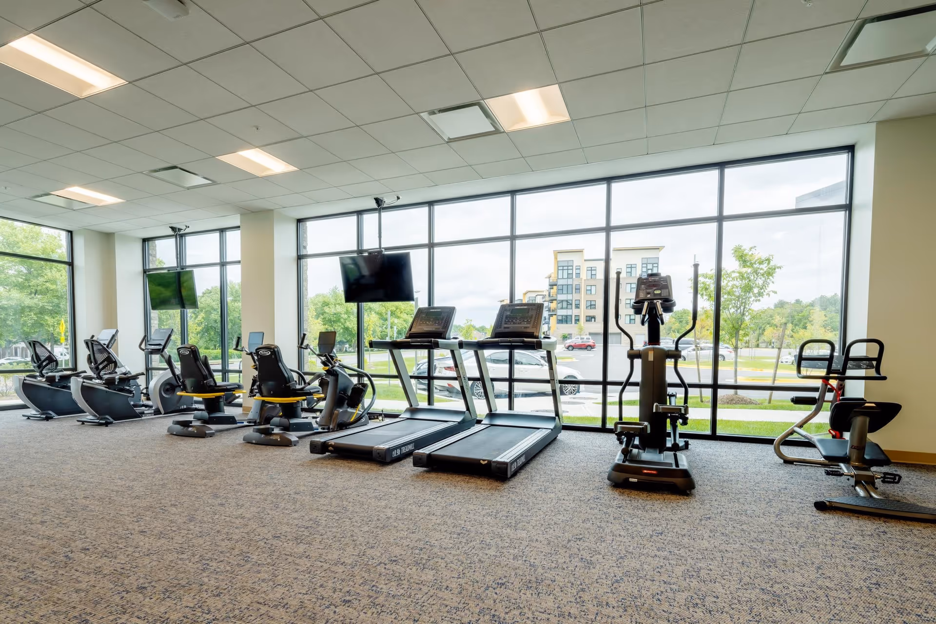 A bright fitness room with large floor-to-ceiling windows showing an outside view of trees, cars, and a building. The room contains several exercise machines including treadmills, stationary bikes, and elliptical trainers, all arranged in a row facing the windows. Two flat-screen TVs are mounted on the walls.
