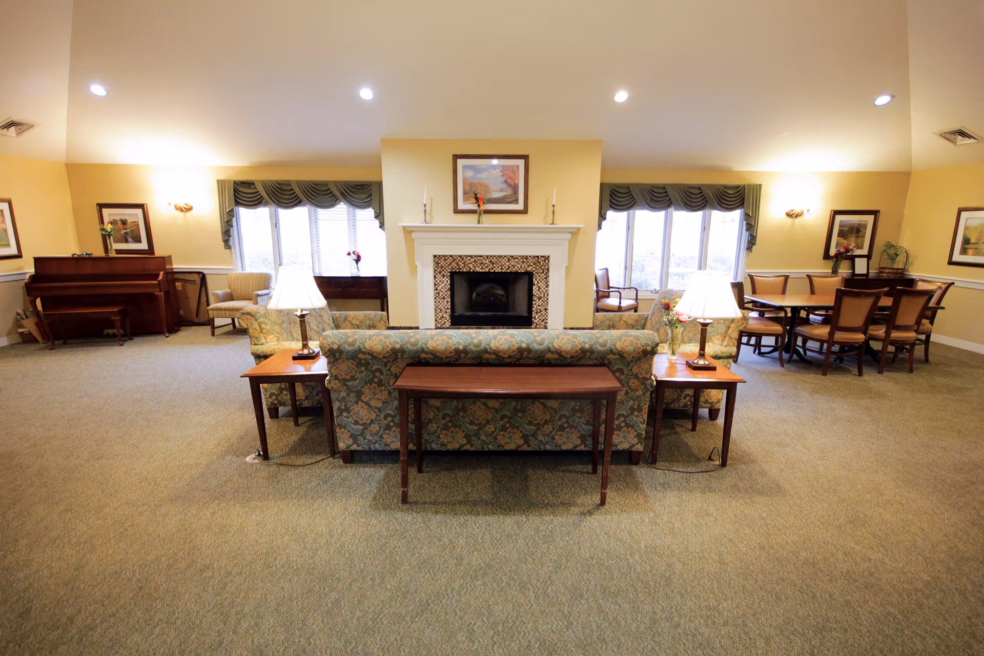 Spacious senior living common room with sofas and side tables facing a fireplace, with a piano and dining table in the background.