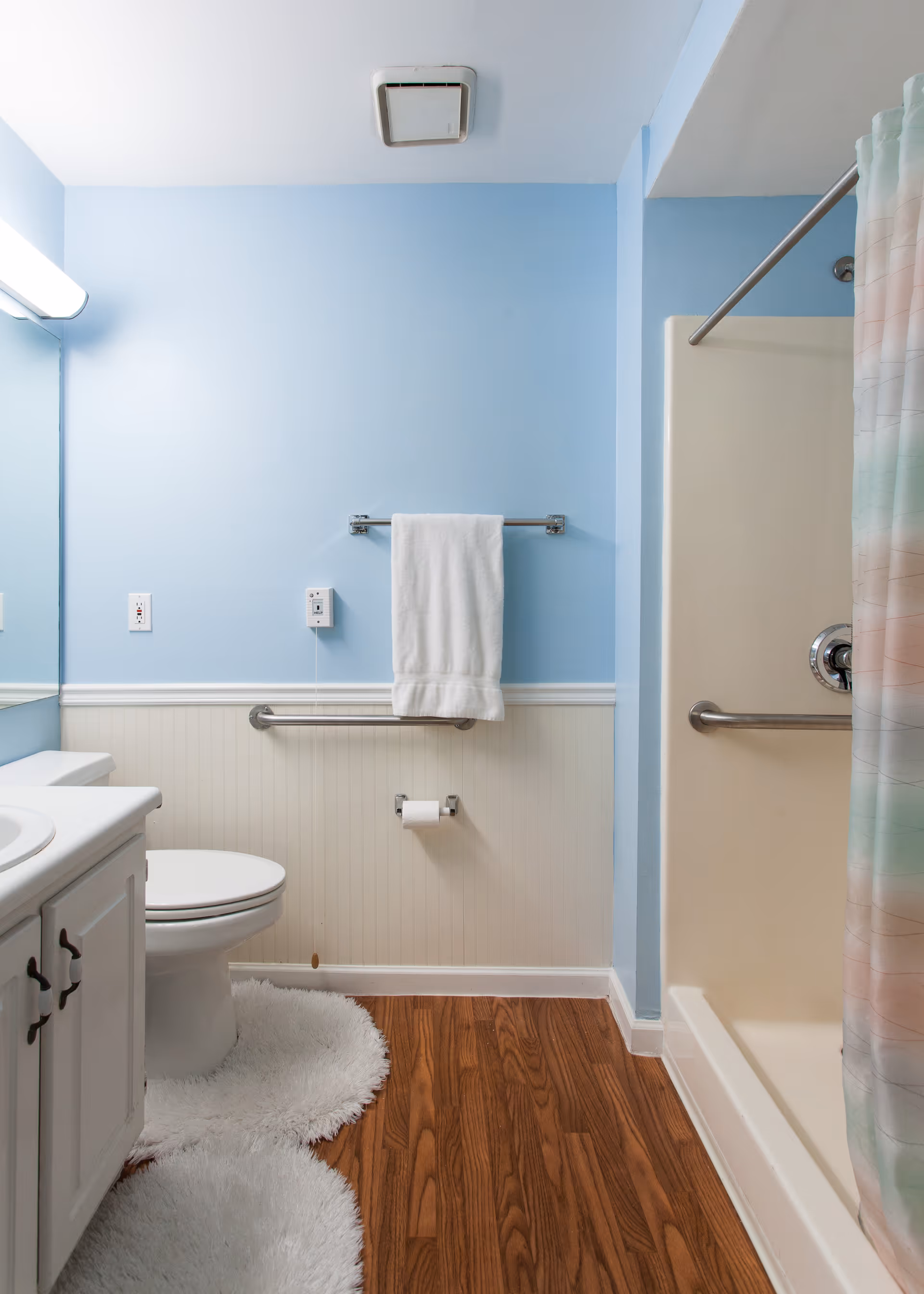 A clean bathroom with light blue walls and white wainscoting. It features a white toilet with a fluffy white rug in front, a white vanity with a sink, a towel rack with a white towel, a toilet paper holder, and a shower with a colorful pastel shower curtain. The floor is wood-patterned, and there is a ceiling vent above.