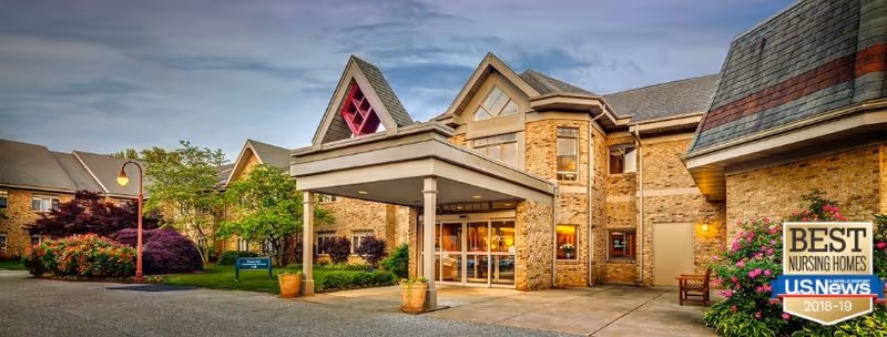 Exterior view of a brick nursing home building with a covered entrance, landscaped greenery, and a clear sky. A sign near the entrance reads 'Best Nursing Homes U.S. News 2018-19'.