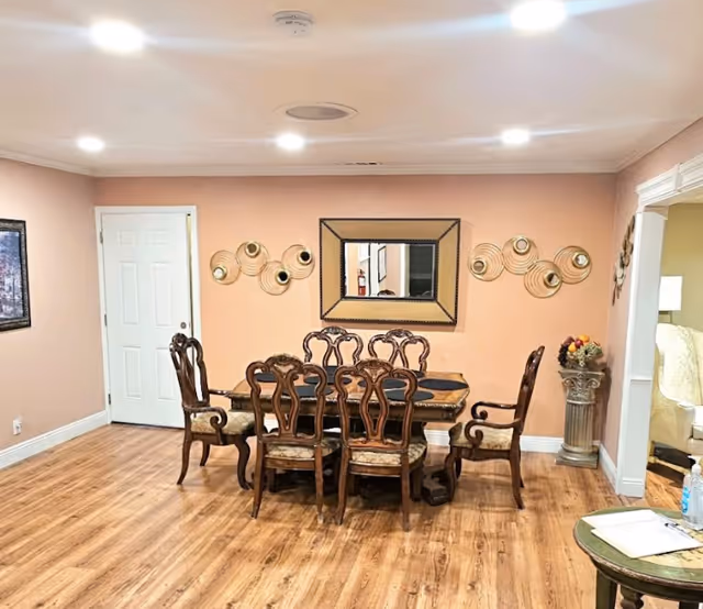 Dining room with a wooden table and six ornate chairs on hardwood floors, peach-colored walls with a mirror and decorative wall art.