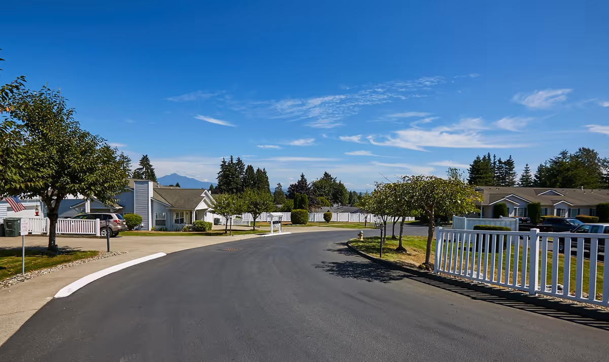A paved road curves through a senior living community with single-story houses on either side, white fences, small trees, and parked cars under a clear blue sky with scattered clouds.