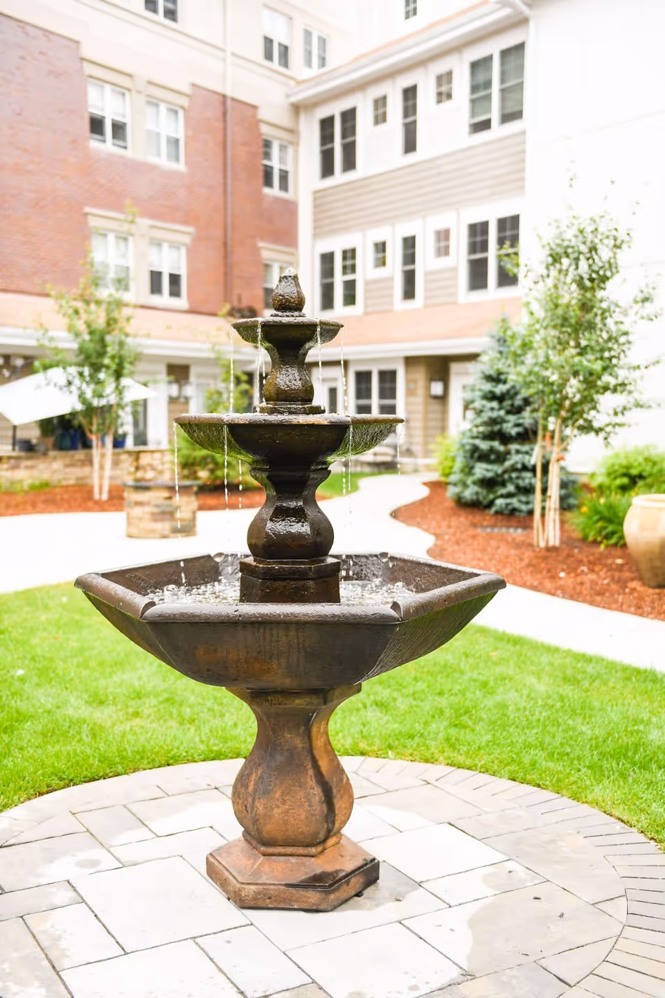 A three-tiered water fountain made of dark stone is situated in a courtyard with green grass, paved walkways, and landscaped plants. Surrounding the courtyard are multi-story buildings with brick and siding exteriors and numerous windows.