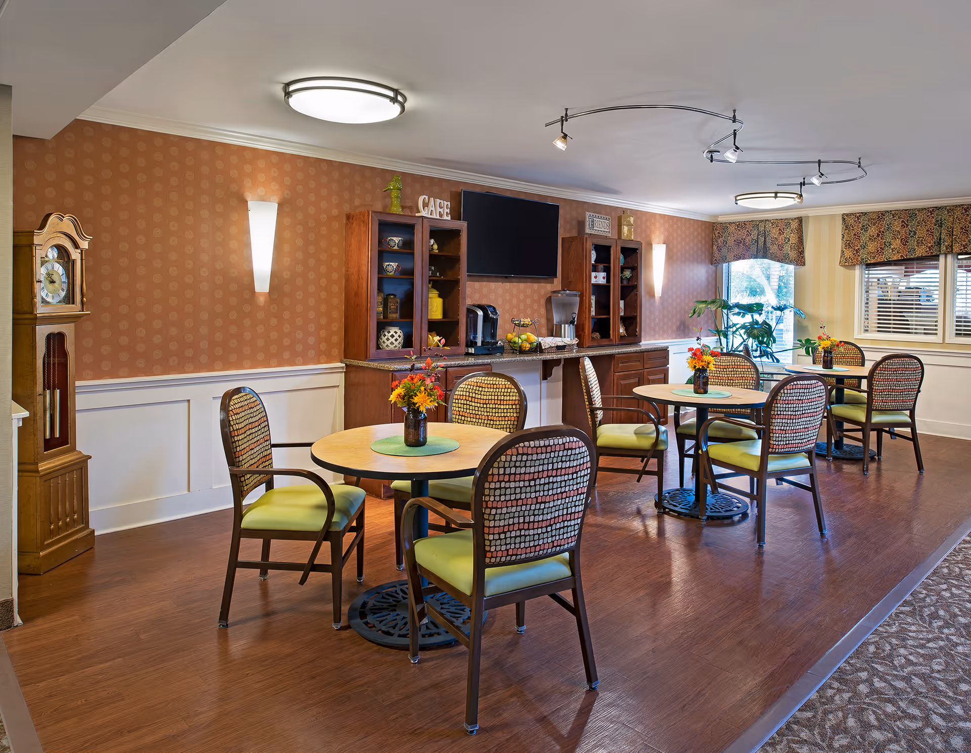 Bright communal dining area with round tables and chairs, floral centerpieces, a coffee station and wall-mounted TV.