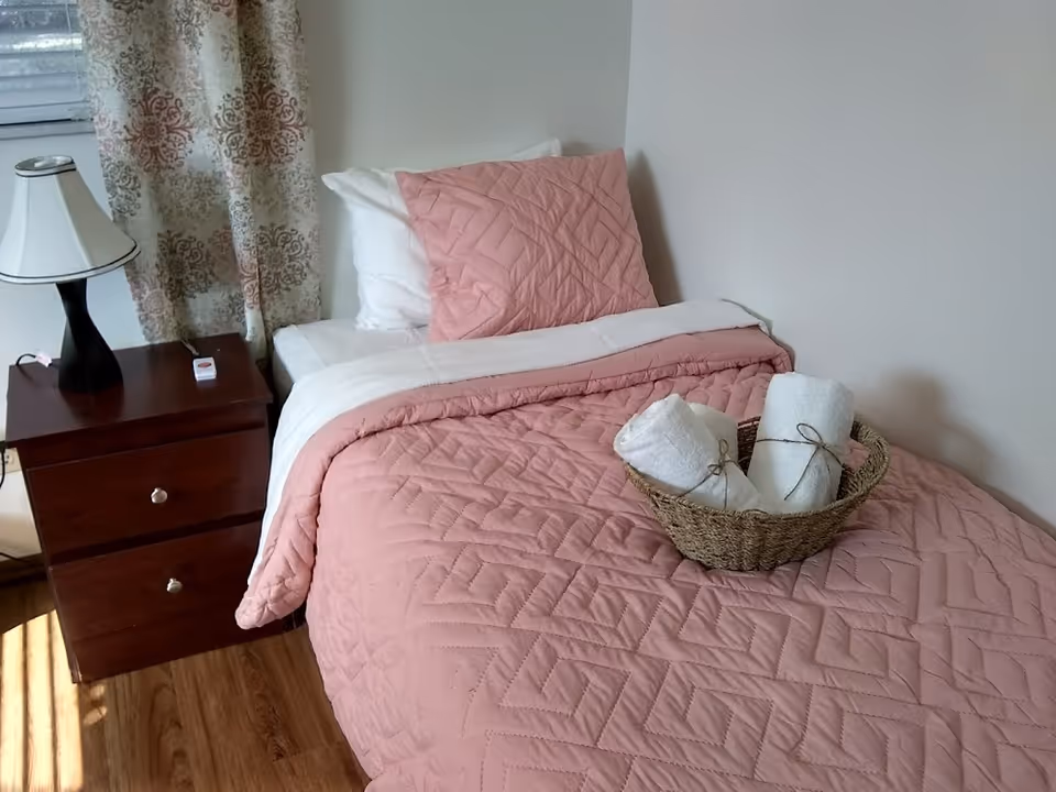 A neatly made single bed with a pink quilted bedspread and matching pillow. On the bed is a woven basket holding two rolled white towels tied with string. Next to the bed is a wooden nightstand with two drawers, a black and white table lamp, and a small white device. A window with patterned curtains is partially visible behind the nightstand.