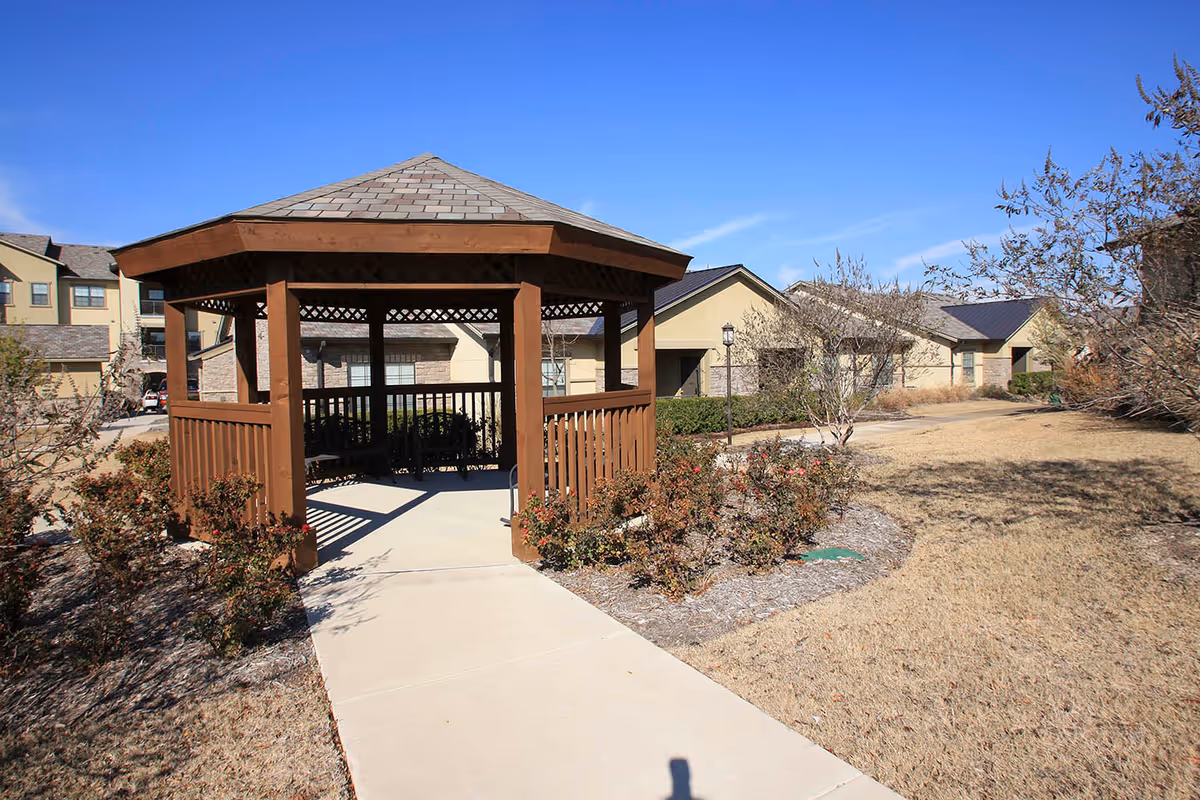 A wooden gazebo with a shingled roof situated on a concrete pathway surrounded by landscaped bushes and dry grass. In the background, there are beige buildings with dark roofs under a clear blue sky.