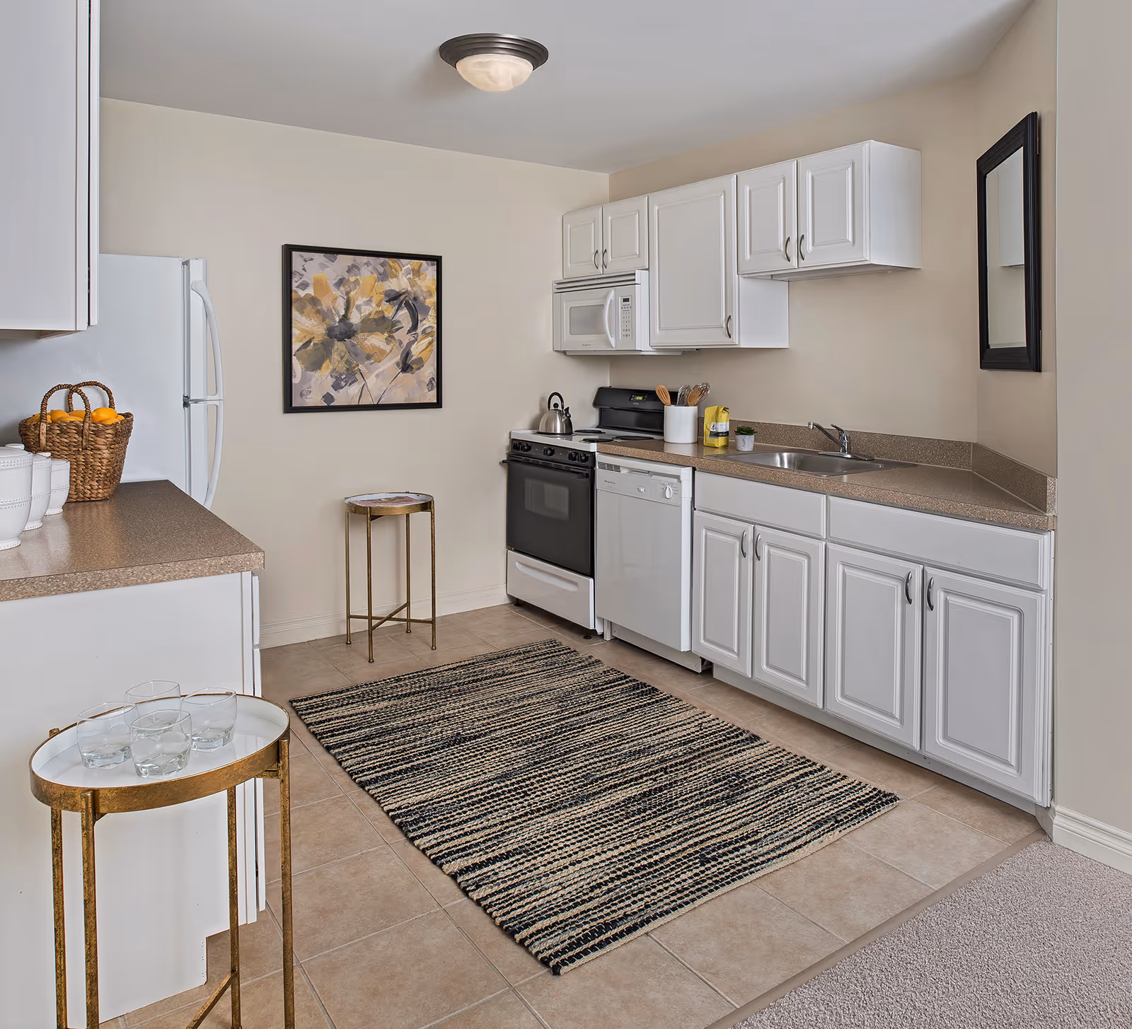 A clean, modern kitchen with white cabinets, a black stove, white microwave, white dishwasher, and a double sink. The floor is tiled with a striped rug in front of the cabinets. There is a small round side table with glasses on it and a basket with oranges on the counter. A framed abstract floral painting hangs on the wall, and a rectangular mirror is mounted on the adjacent wall.