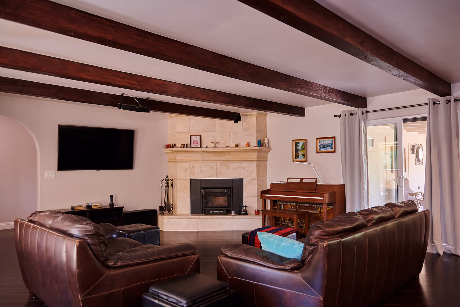 Spacious living room with exposed wooden ceiling beams, leather sofas facing a fireplace and wall-mounted TV, and a piano by sliding glass doors.