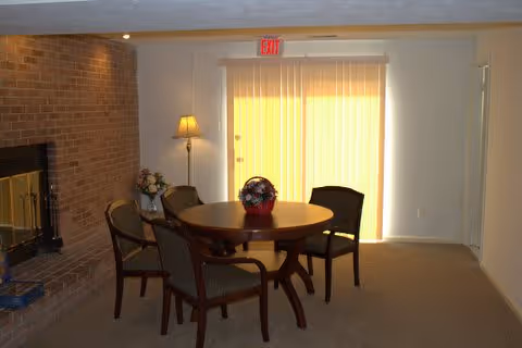 Small common room with a round wooden table and four chairs, a vase of flowers, a floor lamp, a brick fireplace, and a sliding glass door with vertical blinds and an EXIT sign.