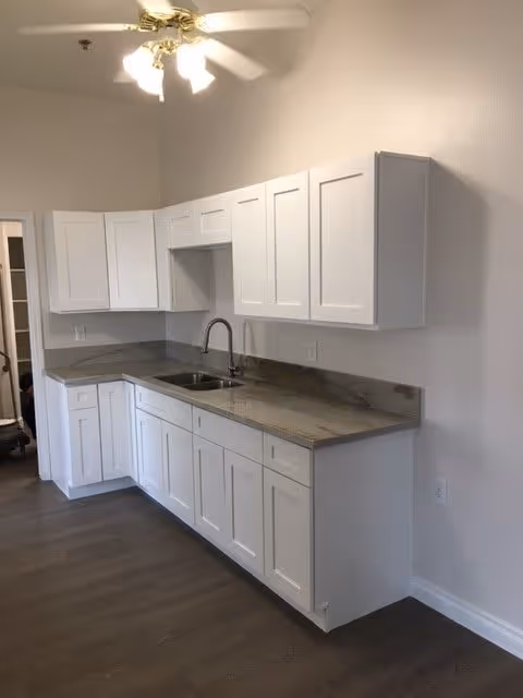 A clean, modern kitchen area with white cabinets and drawers, a marble countertop, and a stainless steel sink with a faucet. The kitchen has a ceiling fan with lights and wood flooring.