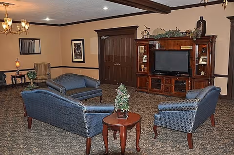 A cozy living room area with three blue upholstered sofas arranged around a small wooden table with a small decorative plant. The room features a large wooden entertainment center with a flat-screen TV, decorative items on top, and framed artwork on the walls. The carpet has a patterned design, and the walls are painted beige with dark wood trim.