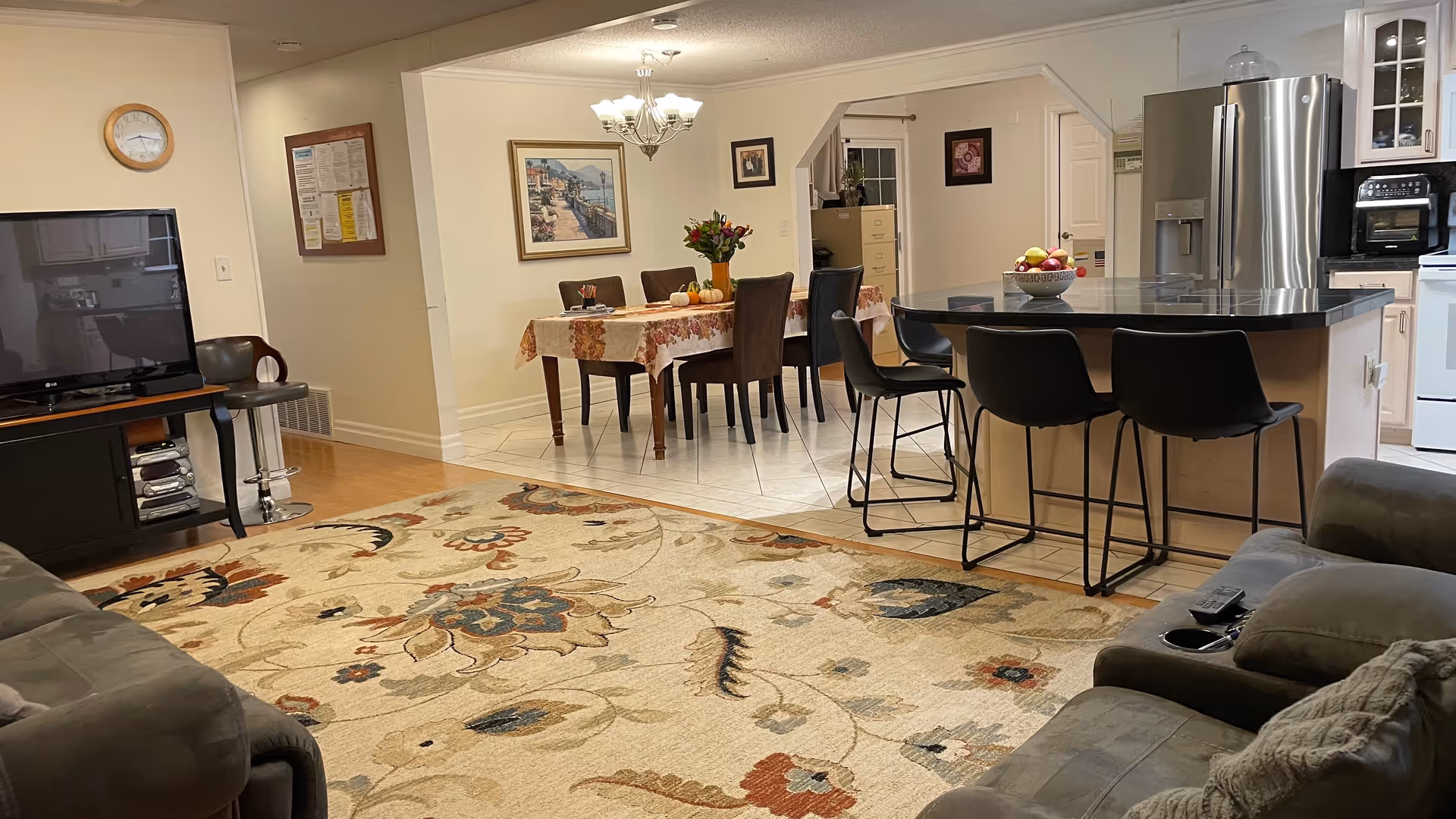 Open living area with a sofa and TV in the foreground, a dining table in the middle, and a kitchen island with bar stools to the right.