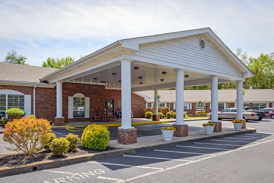 Entrance of Life Care Center of Centerville showing a covered drop-off area with white columns and brick bases, surrounded by landscaped bushes and a parking lot with several cars.