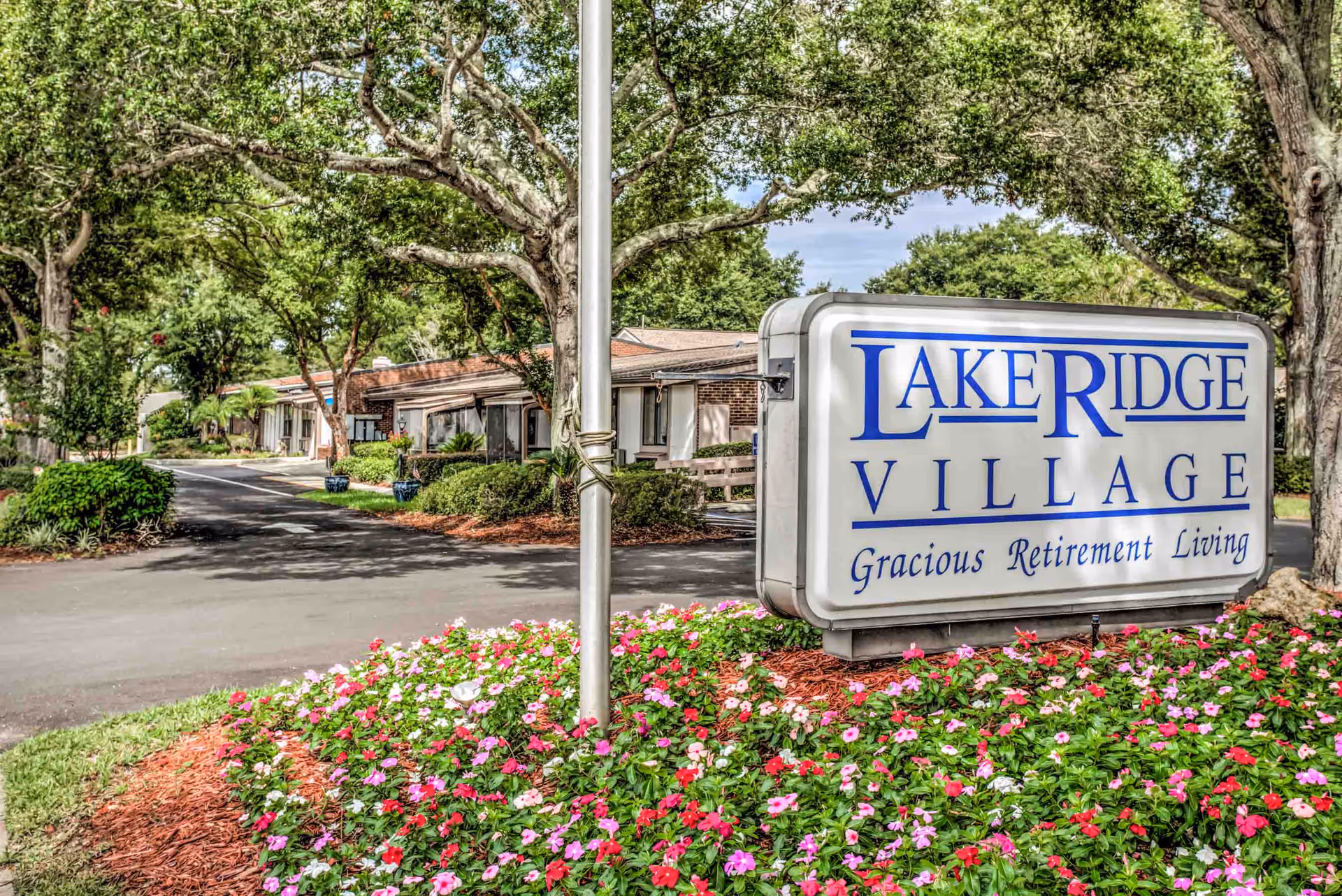 Entrance sign for Lake Ridge Village, a gracious retirement living community, surrounded by colorful flowers and trees with a driveway and single-story buildings in the background.