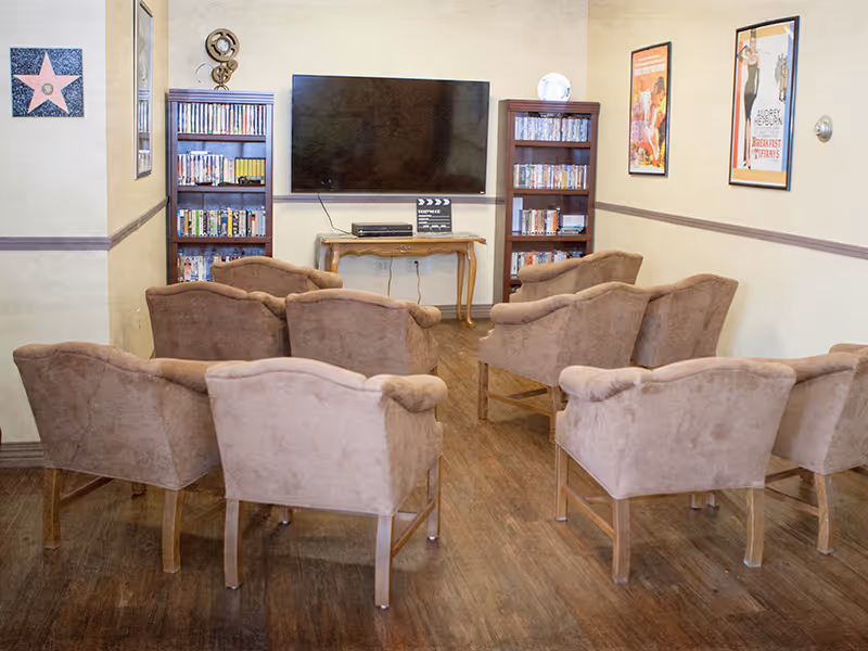 A cozy media room with eight brown upholstered chairs arranged in two rows facing a large flat-screen TV mounted on a beige wall. Below the TV is a small wooden table with a DVD player and a clapperboard. On either side of the TV are tall bookshelves filled with DVDs. The room has wooden flooring and framed movie posters on the walls.