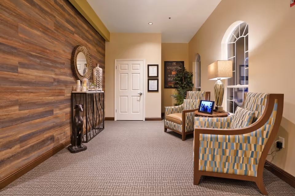 A cozy indoor seating area with two patterned armchairs flanking a small table holding a lamp and tablet, opposite a wood-accent wall with a console table and decorative mirror.